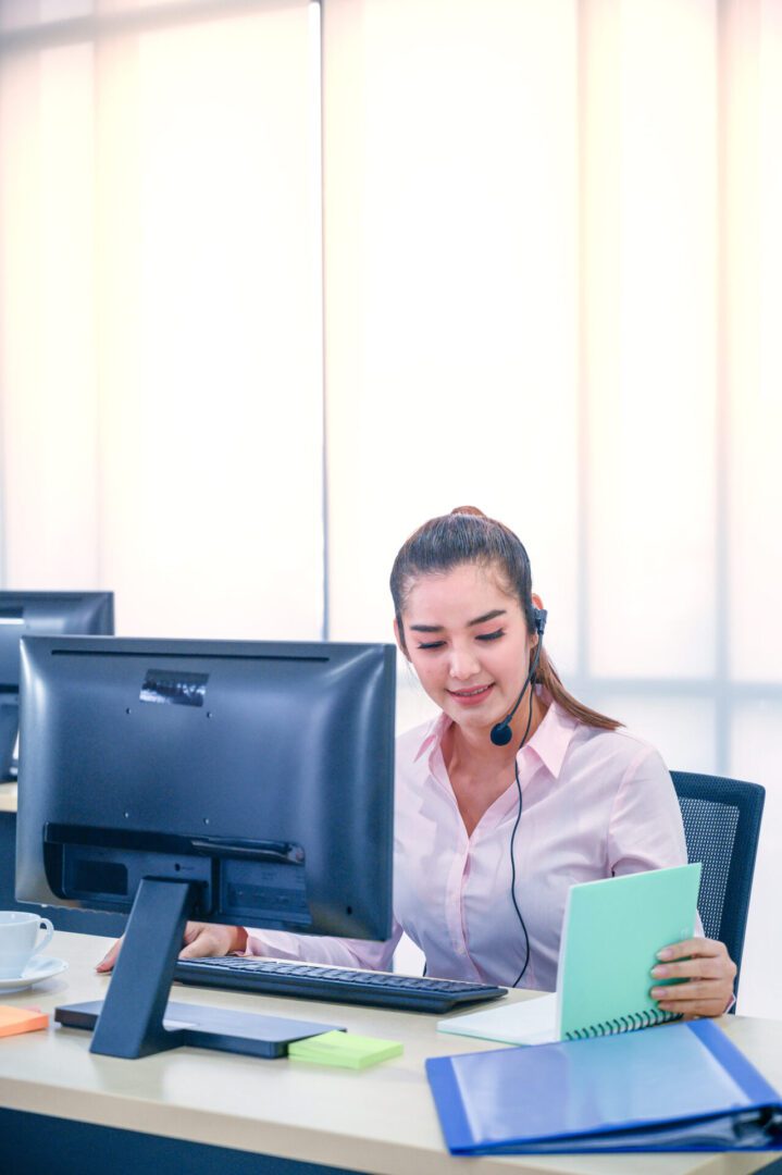 A woman with a headset working at a computer, holding a phone.